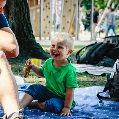 Wir sehen einnen Jungen, auf einer Picknickdecke sitzen. Er lacht und hält eine Brezel in der Hand.