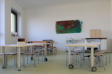 Auf dem Foto ist das Atelier zu sehen. An der großen Wand hängt ein selbstgestaltetes Bild und davor stehen zwei große Tische mit Malmaterialien.