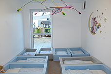 Ein heller Schlafraum mit vielen Betten und einem großen Fenster. An der Wand ist ein Wandtattoo und von der Decke hängt eine Spieluhr 