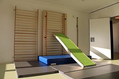 Auf dem Foto ist eine Turnhalle zu sehen. Eine große Sprossenwand, an der eine Rutsche eingehangen ist. Unter dieser befindet sich eine große Turnmatte.